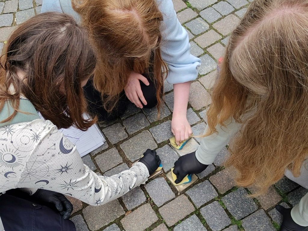 Drei Schülerinnen reinigen in der Bünder Innenstadt gemeinsam Stolpersteine im Pflaster. Mit Schwämmen und Schutzhandschuhen säubern sie die Messingplatten, um an die Opfer des Nationalsozialismus zu erinnern.