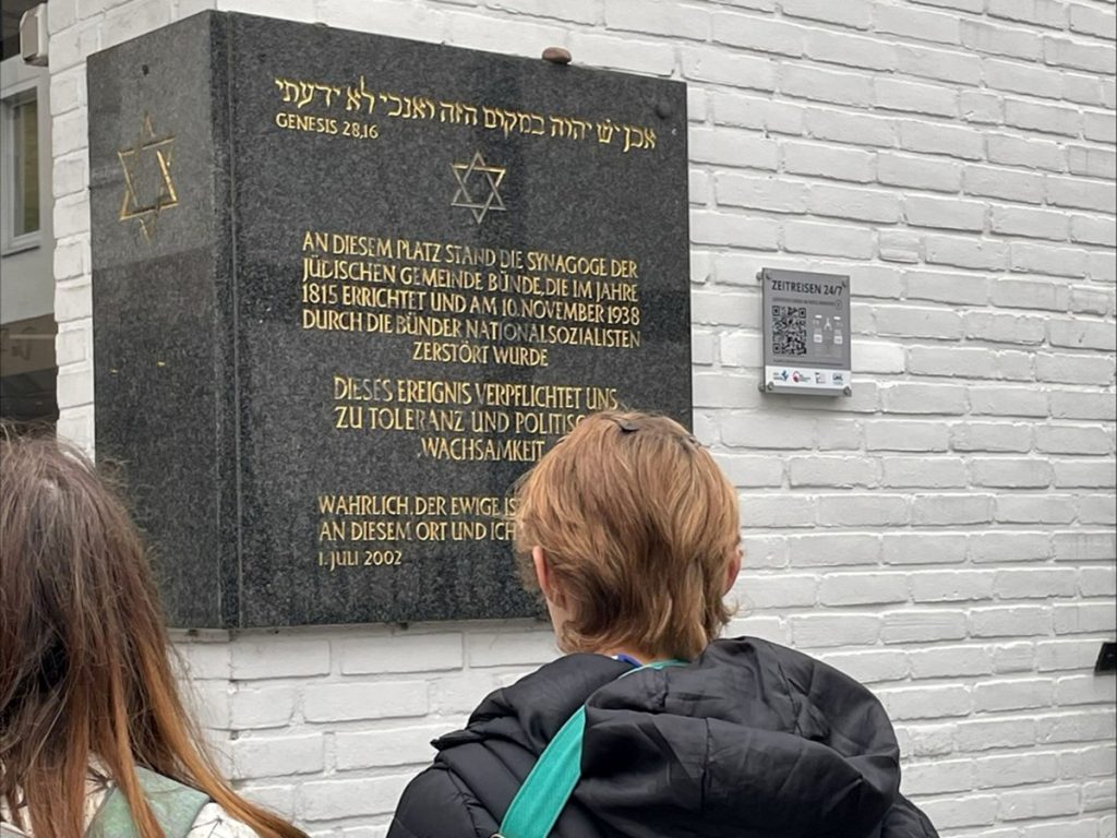 Schüler:innen stehen vor der Gedenktafel am ehemaligen Standort der Synagoge in Bünde. Die Tafel erinnert an die jüdische Gemeinde und an die Zerstörung der Synagoge während der Novemberpogrome 1938.