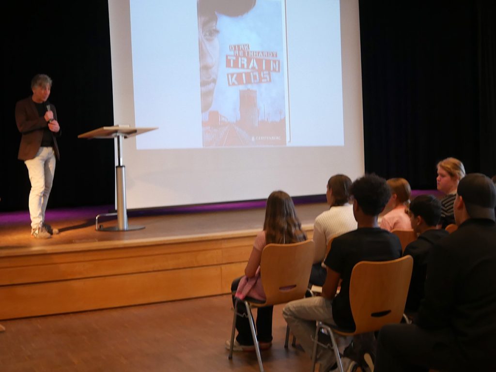 Der Jugendbuchautor Dirk Reinhardt liest vor Schüler:innen aus seinem Roman „Train Kids“. Die Lesung findet in der Aula statt und wird durch eine Projektion des Buchcovers begleitet.