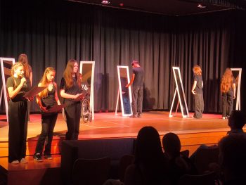 Schüler:innen der Wahlpflichtkurse Darstellen und Gestalten präsentieren auf der Bühne der Aula eine Szene aus dem Programm „Im Spiegel“. Mehrere Spiegelrequisiten und szenische Lesungen greifen das Leitmotiv des Abends auf.