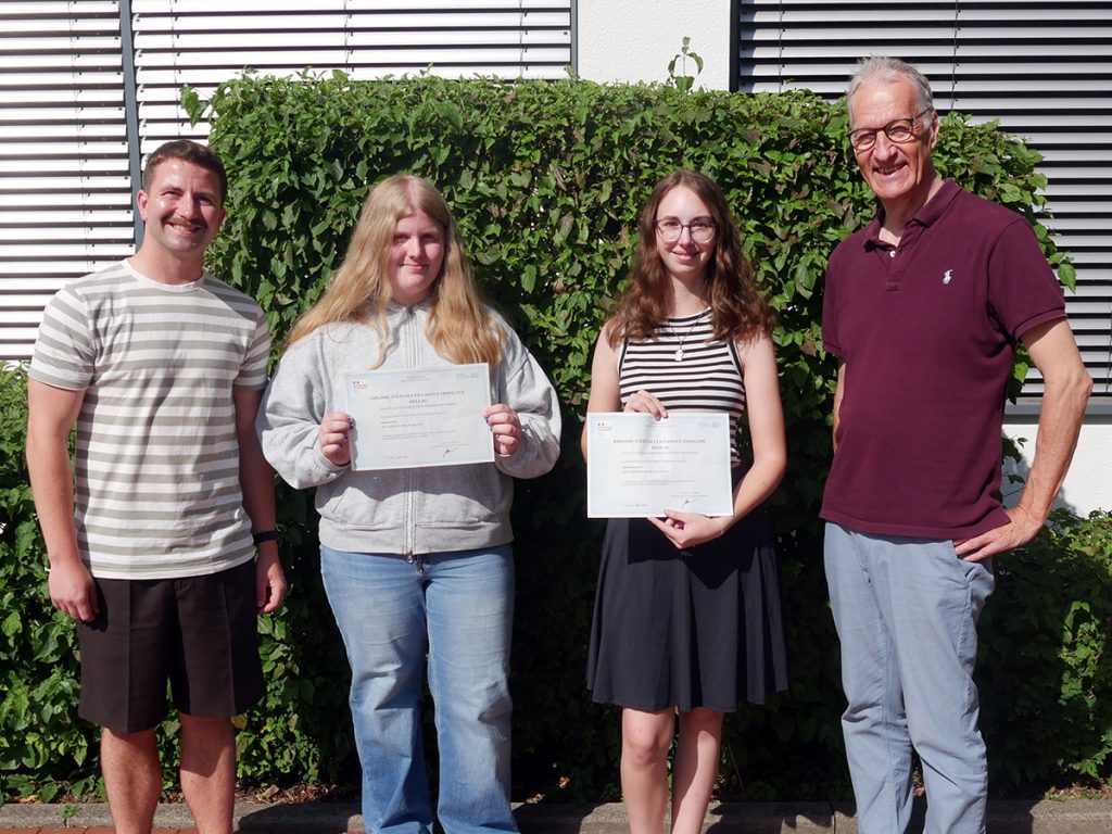 Zwei Schülerinnen präsentieren gemeinsam mit Französischlehrer Matthias Janoschka und Schulleiter Andreas Hillebrand ihre DELF-Diplome. Die Auszeichnungen bescheinigen erfolgreich erworbene Französischkenntnisse auf unterschiedlichen Niveaustufen.