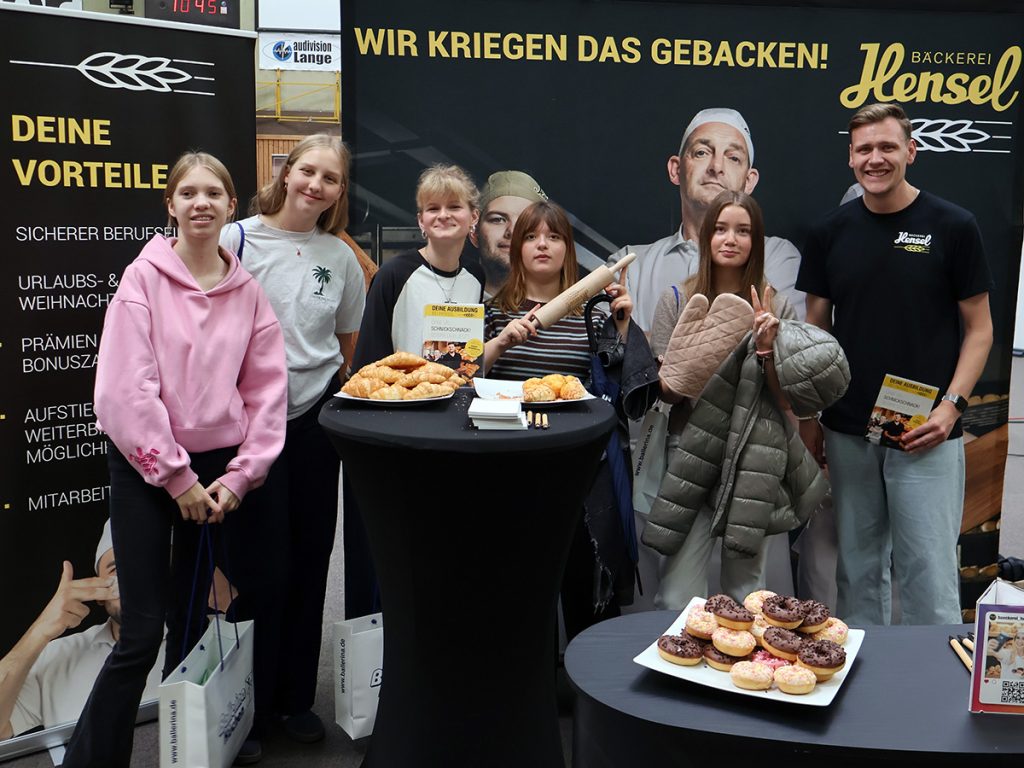 Fünf Schülerinnen der Gesamtschule Rödinghausen stehen gemeinsam mit einem Mitarbeiter am Messestand der Bäckerei Hensel. Auf den Tischen liegen verschiedene Backwaren und Informationsmaterialien.