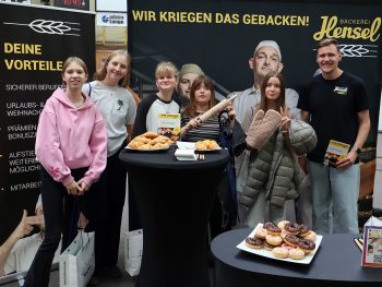 Fünf Schülerinnen der Gesamtschule Rödinghausen stehen gemeinsam mit einem Mitarbeiter am Messestand der Bäckerei Hensel. Auf den Tischen liegen verschiedene Backwaren und Informationsmaterialien.